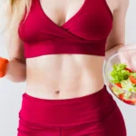 Fit woman in red sportswear holding a dumbbell and salad, promoting healthy lifestyle.