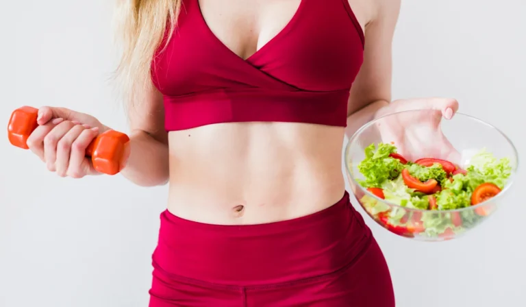 Fit woman in red sportswear holding a dumbbell and salad, promoting healthy lifestyle.