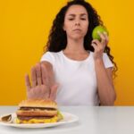 A woman in a white shirt holds a burger with a thoughtful expression. She's against a bright yellow background, creating a vibrant, lively atmosphere.
