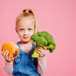 A child in denim overalls holds a green toy dinosaur and an orange toy, against a pink background. They are smiling with a playful expression.