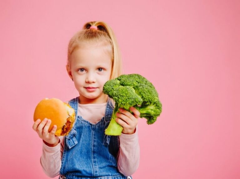 A child in denim overalls holds a green toy dinosaur and an orange toy, against a pink background. They are smiling with a playful expression.