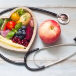 A fresh fruit bowl containing bananas, apples, and berries is displayed next to a stethoscope and a blood pressure cuff, symbolizing health and wellness.
