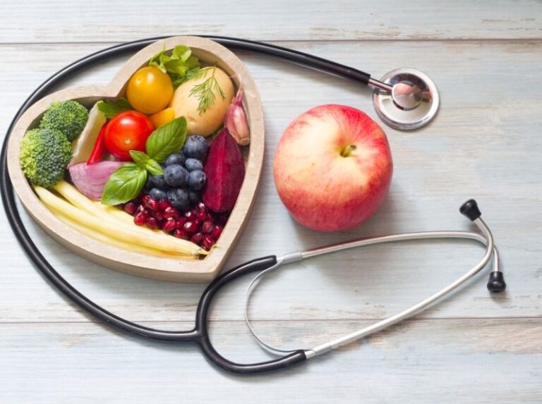 A fresh fruit bowl containing bananas, apples, and berries is displayed next to a stethoscope and a blood pressure cuff, symbolizing health and wellness.