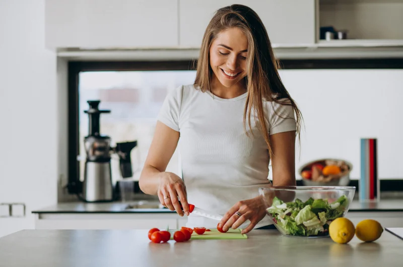 Smiling woman in kitchen holding a tablet, surrounded by vegetables and cooking utensils, creating a warm and inviting atmosphere focused on cooking.